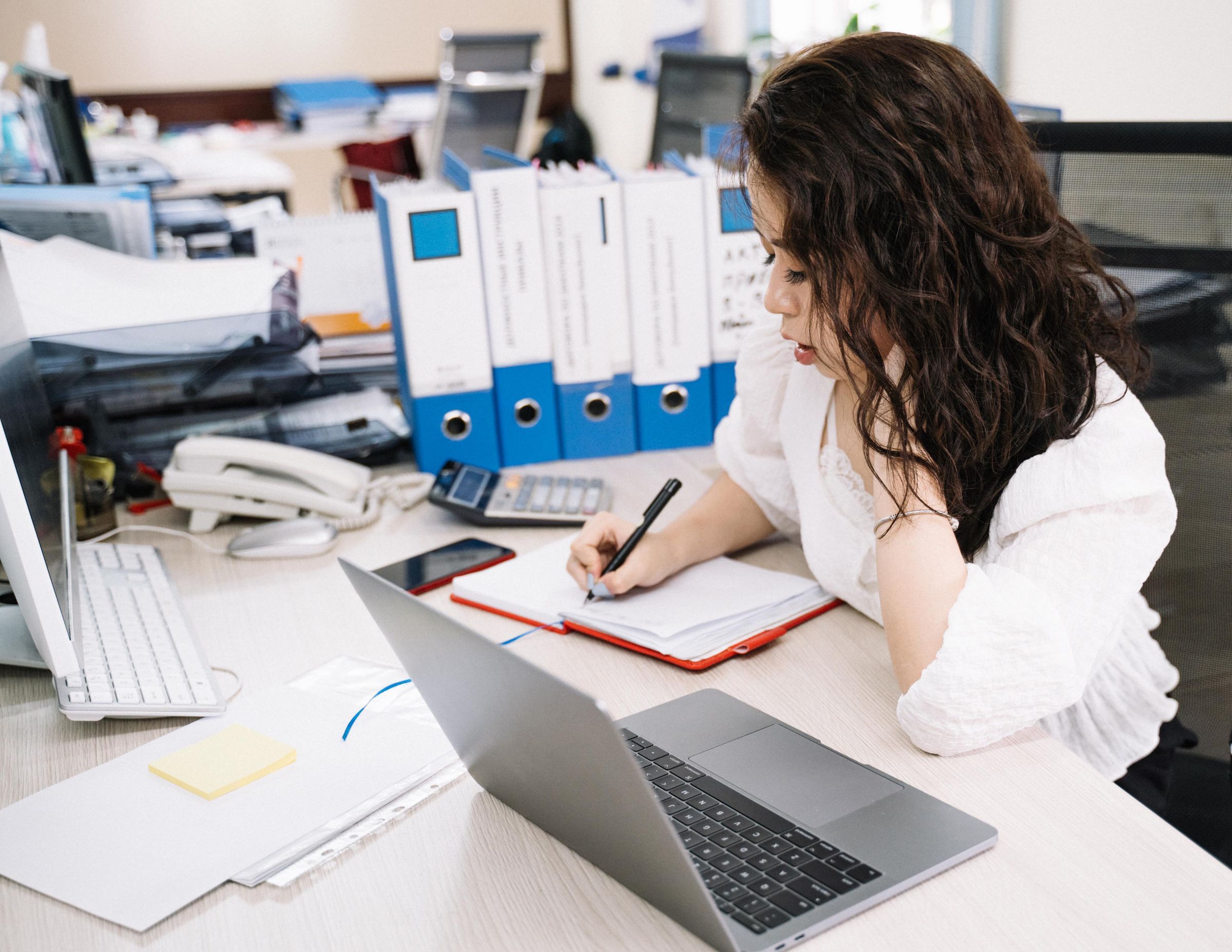 Büro mit Aktenordnern im Regal, eine Mitarbeiterin schreibt am Schreibtisch neben ihrem Laptop.